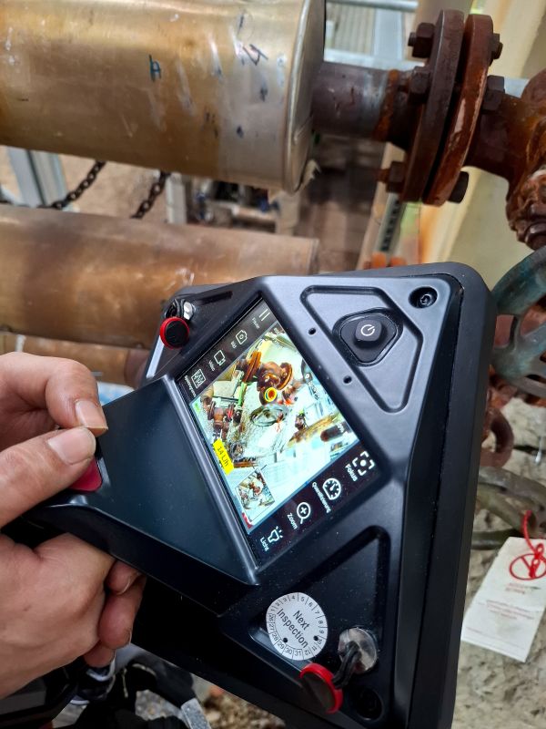 Hand holding an ultrasonic camera in front of a rusty flange connection, display showing the acoustic overlay